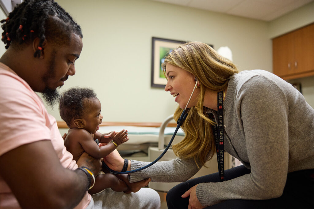Pediatrician using a stethoscope to examine a smiling baby held by a parent during a checkup.