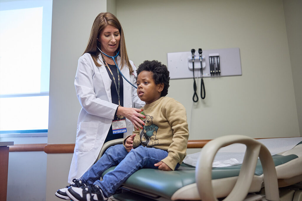 Pediatrician using a stethoscope to examine a young child sitting on an exam table in a medical office.